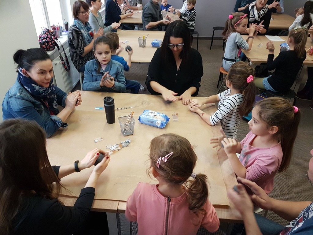 Powiększ obraz: Dorośli i dzieci siedzą razem przy stole i lepią gwizdki z gliny